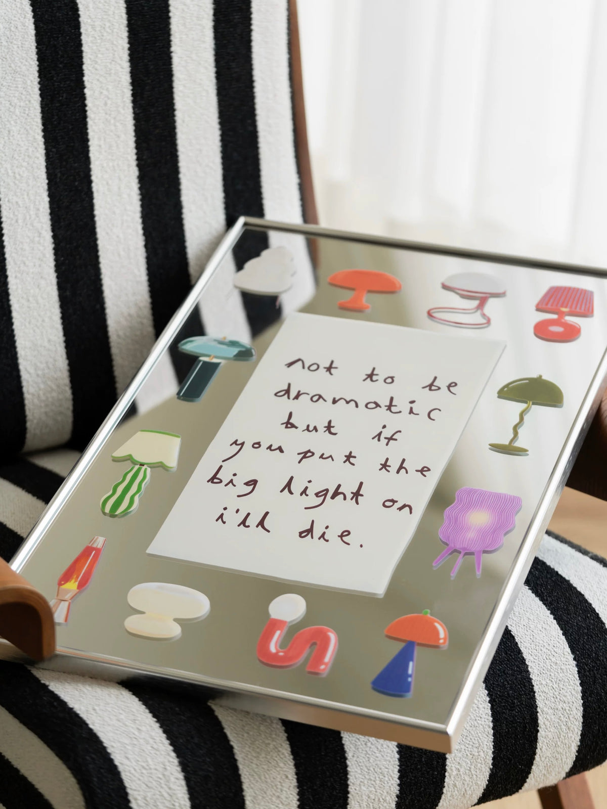 A printed mirror rests at an angle, showing colorful lamp illustrations, handwritten text and a slim silver frame.