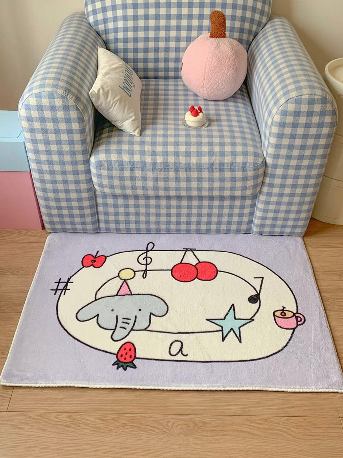 A purple cartoon rug sits in front of a checked armchair, with cherries and an elephant inside a cream oval.