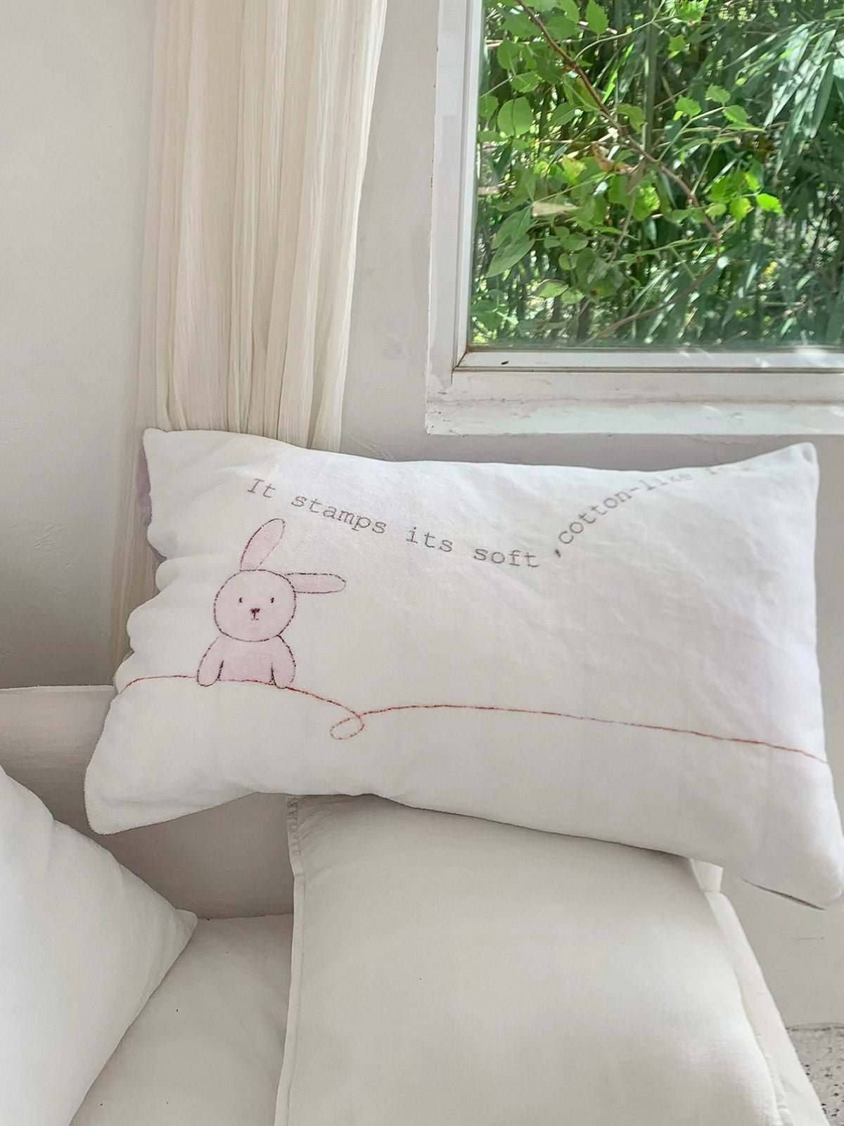A rabbit pillowcase rests on a pale chair beside a window with green foliage.