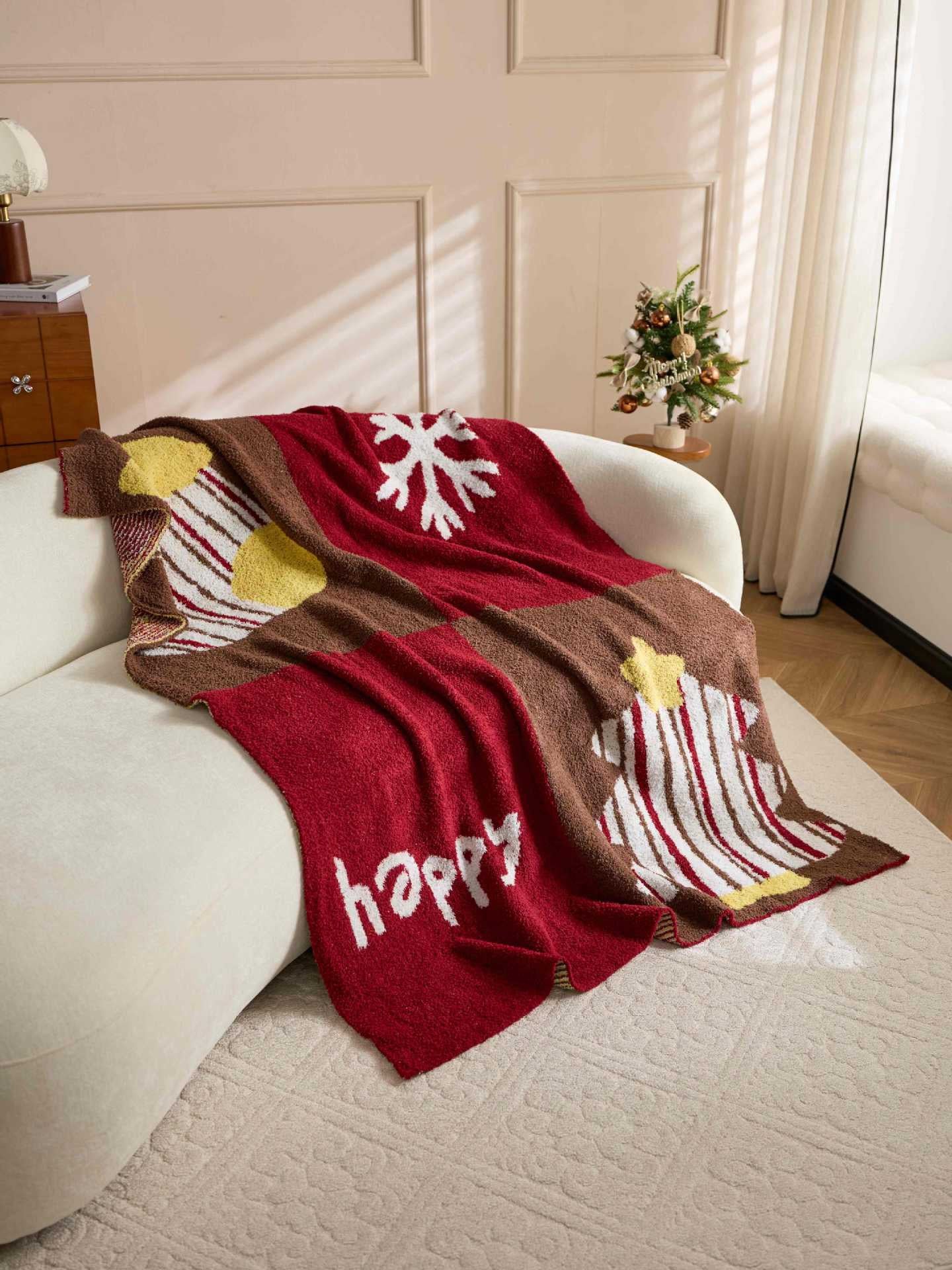 A cozy knitted throw blanket draped over a cream-colored sofa in a sunlit room. The blanket features a holiday-themed pattern with a large white snowflake, a striped Christmas tree, a yellow star, and the word 'happy' in white script on a deep red background.