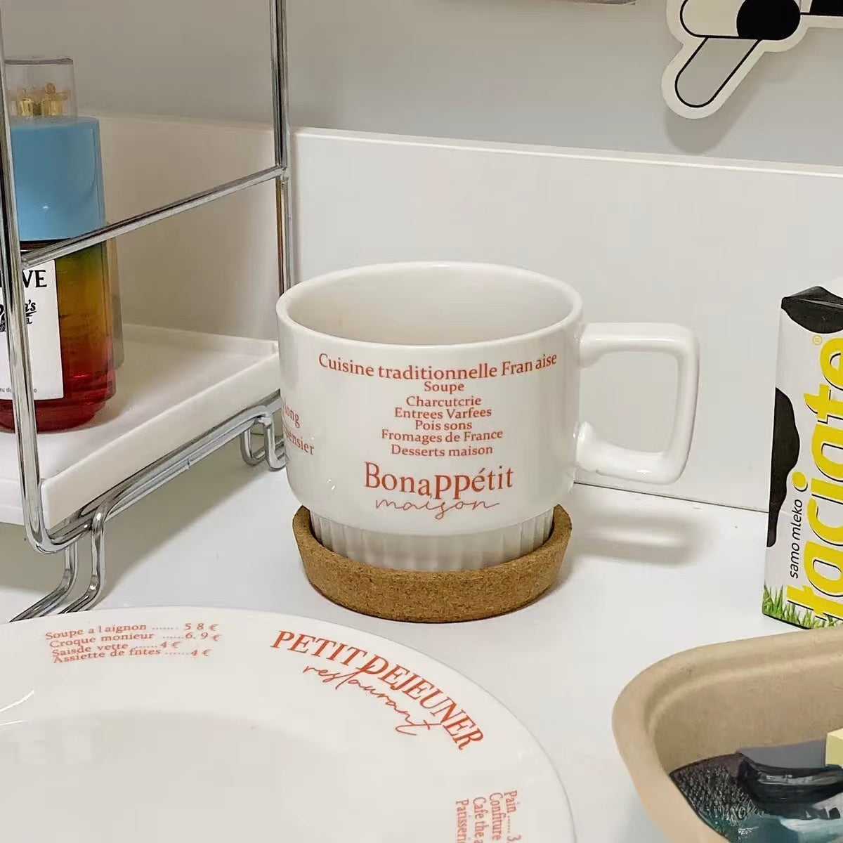 A white ceramic coffee mug with red text reading 'BonaPétit maison' sits on a round cork coaster on a white countertop. The background shows a kitchen-like setting with a metal shelf, a plate, and other household items.