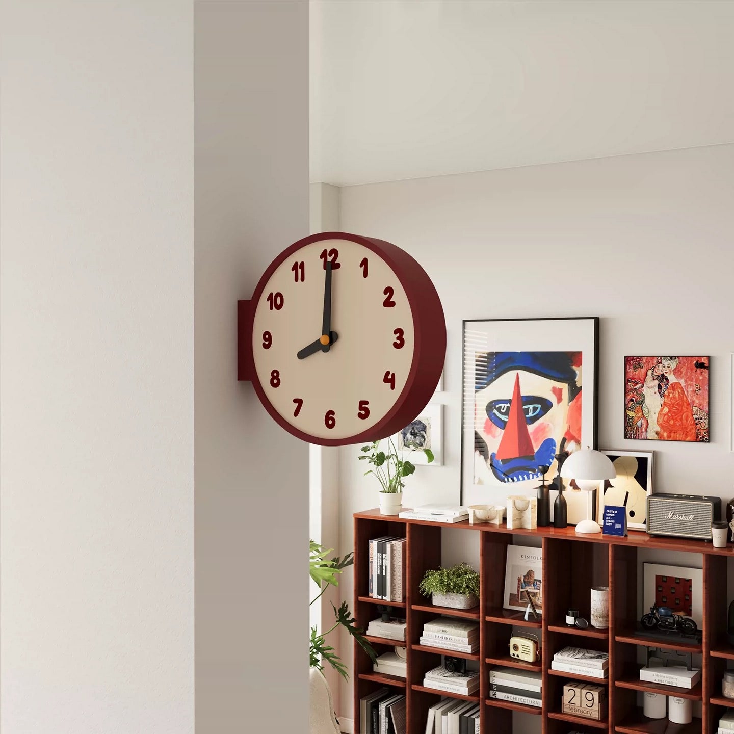 A round wall clock with a cream face, dark red outer rim, and black hands, mounted on a white wall in a styled interior room with bookshelves and framed art.