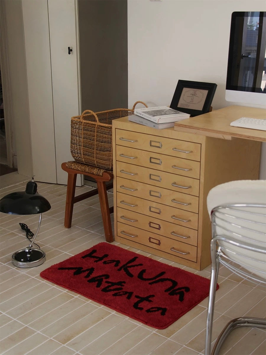 A red rectangular bath mat with black lettering sits below a drawer cabinet in a wider corner view.