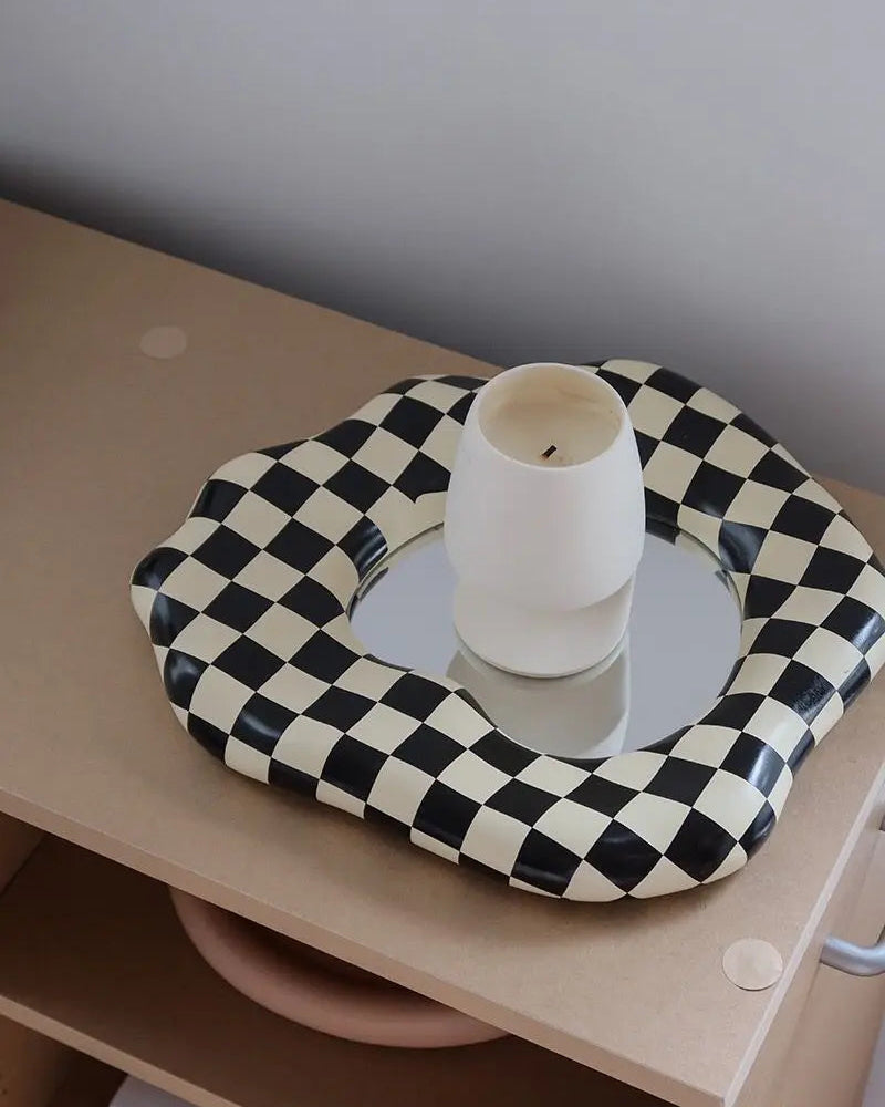 A black and cream checkerboard upholstered mirror tray with a white ceramic candle holder in the center, styled on a light wood shelf against a neutral gray wall.
