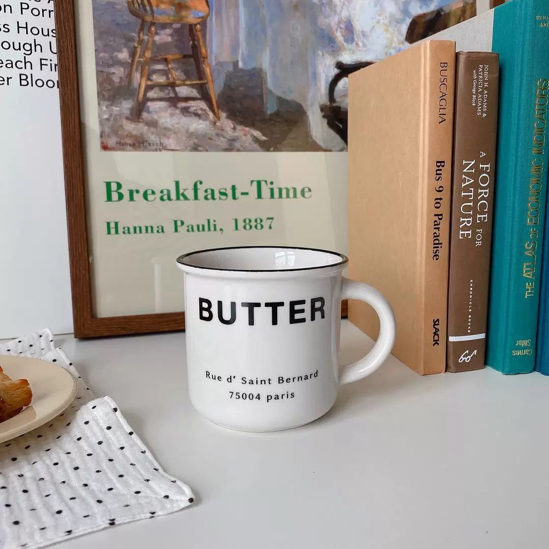 A white ceramic coffee mug with black text reading 'BUTTER' and a street address, styled on a tabletop next to books and a framed print.