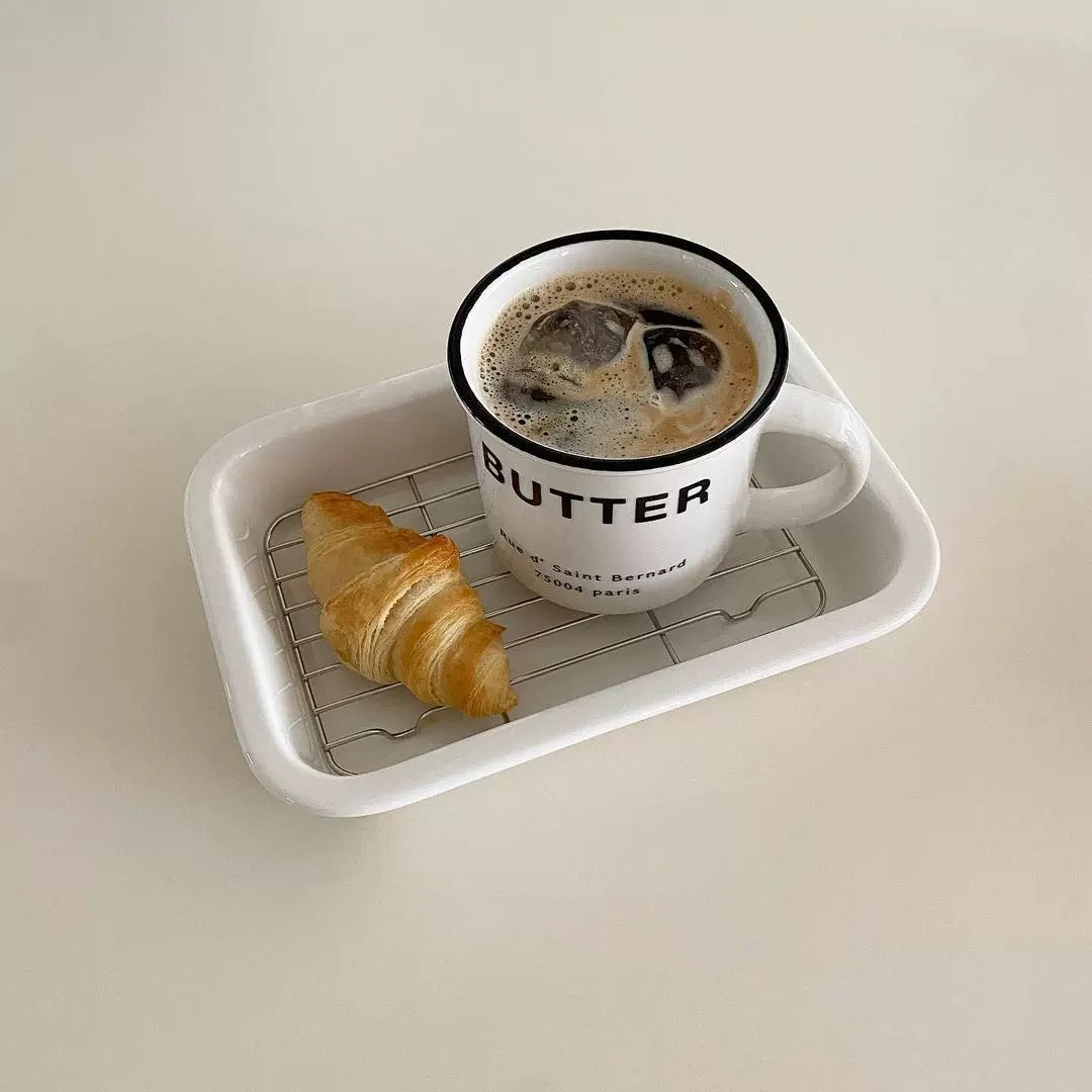 A white rectangular tray with a metal cooling rack, holding a white enamel coffee mug with 'BUTTER' printed on it and a croissant, set against a plain light beige background.