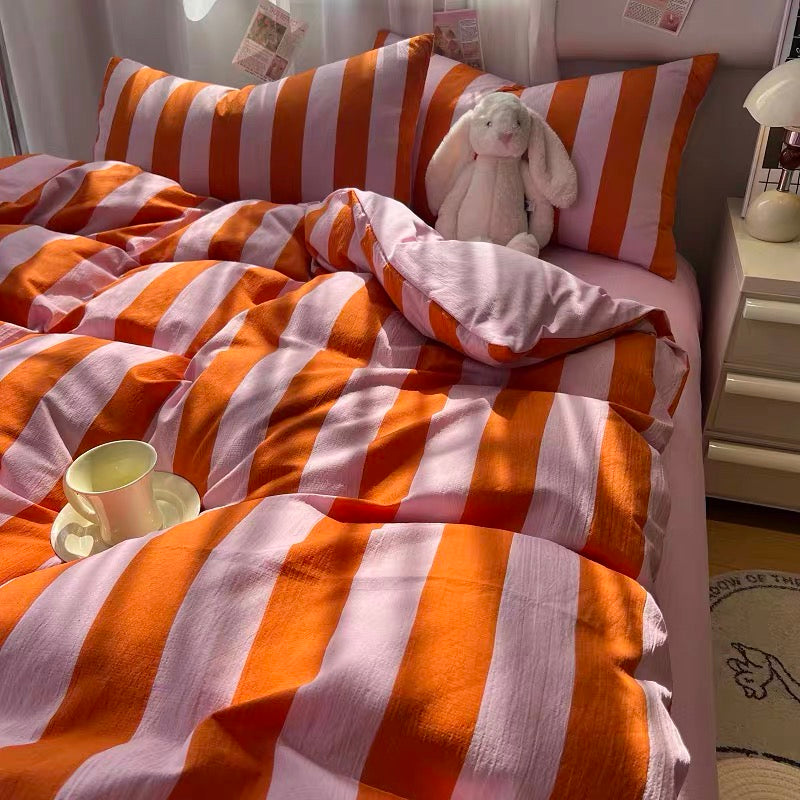 A bed made up with orange and white striped bedding, including a duvet cover and matching pillowcases, with a white plush bunny toy on the pillow. A small cup sits on the bed, and a bedside table and lamp are visible in a softly lit bedroom setting.