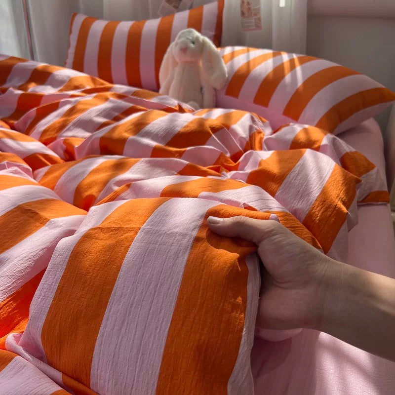 A close-up of a bed with orange and white striped bedding, including a duvet cover and pillowcases, with a hand pulling the duvet to show its soft texture. A white plush bunny toy is resting on the pillows in the background.