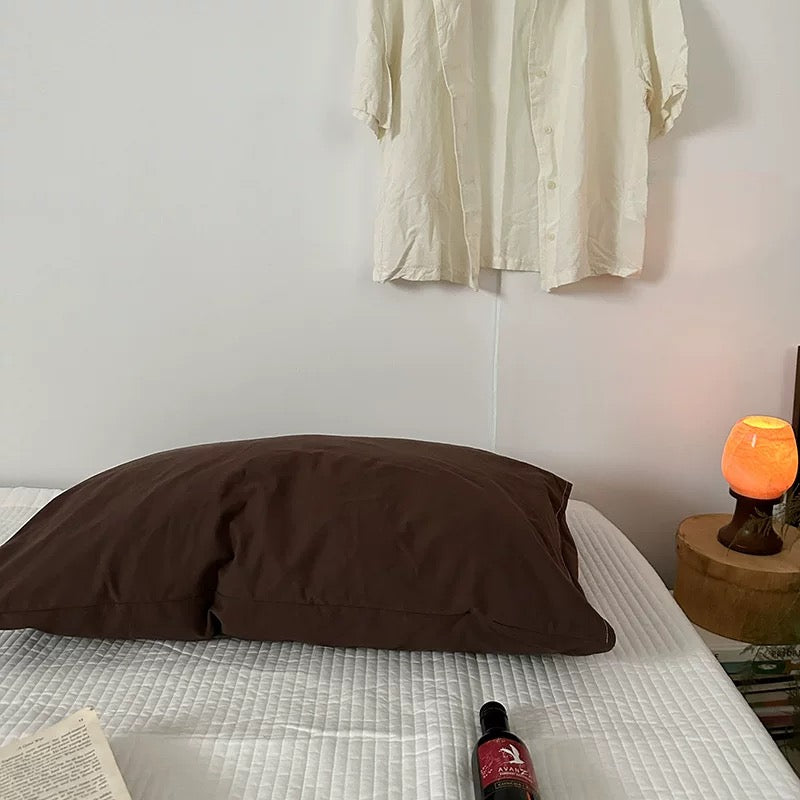 Brown pillow on a bed with a bottle and book, against a plain wall.