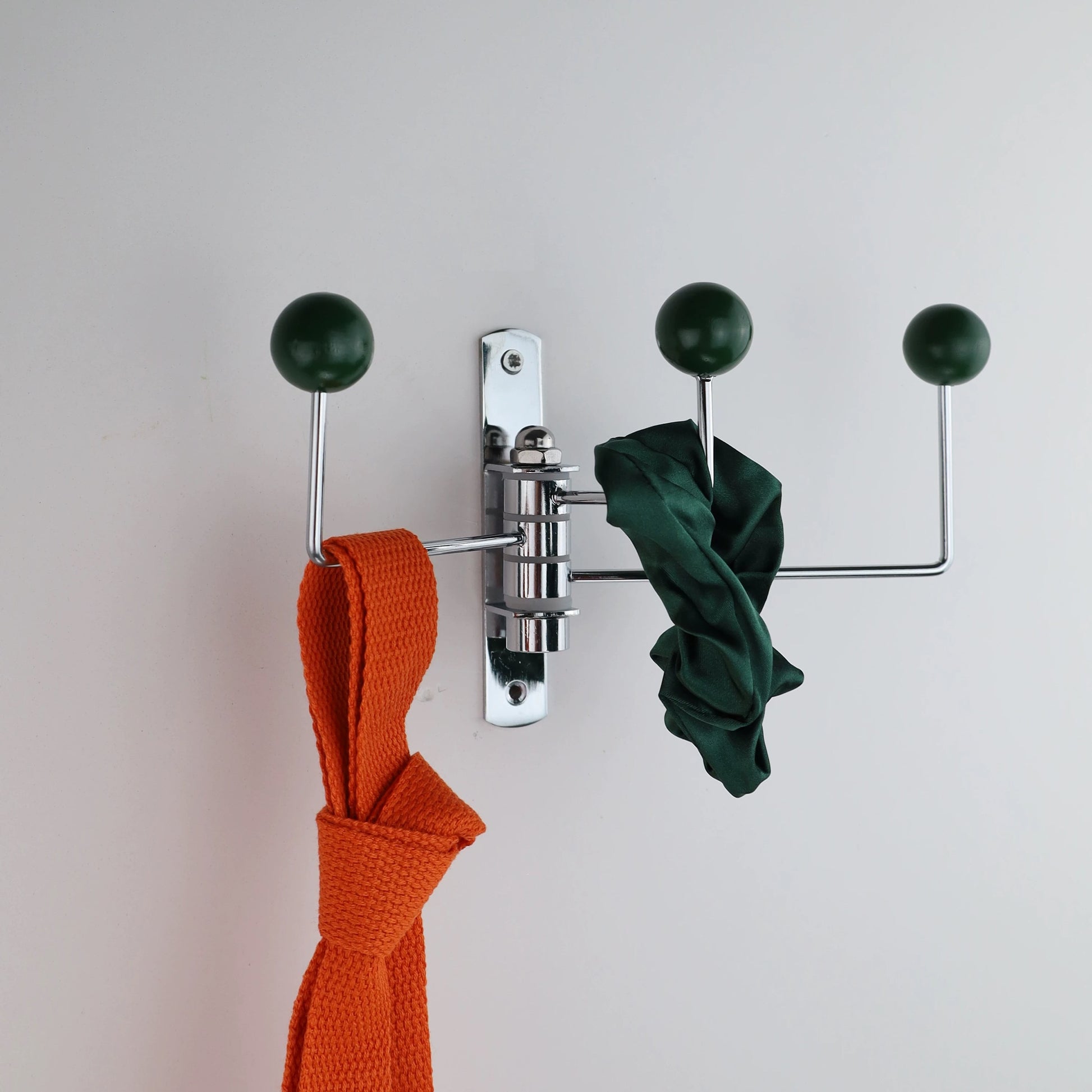 A wall-mounted three-hook coat rack with a polished silver metal frame and dark green spherical hook knobs. An orange woven scarf hangs from the left hook, a dark green fabric item hangs from the center-right horizontal bar, and a third dark green scarf is draped over the right vertical hook, all against a plain light gray wall.
