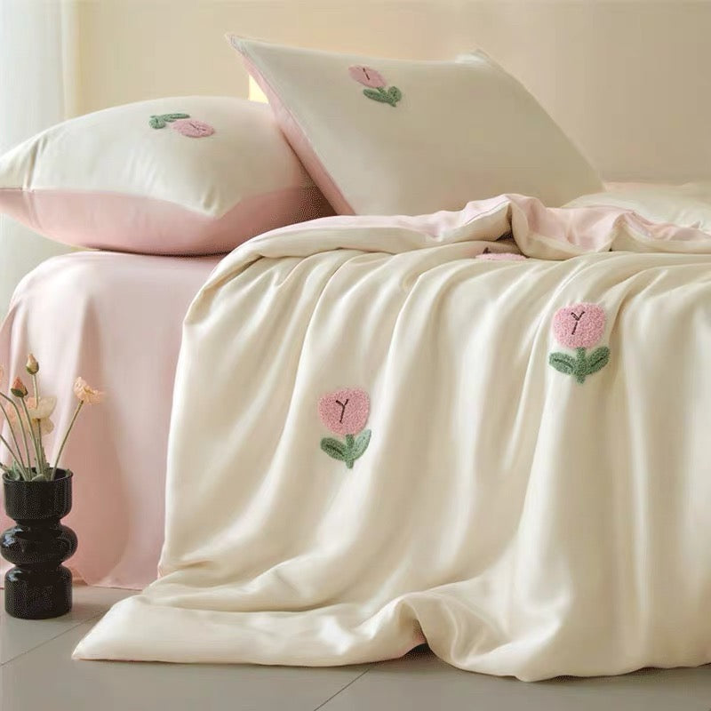 A cream-colored satin throw blanket draped over a bed, decorated with pink and green embroidered flowers. The bed is styled with matching cream and pink pillows, and a small black vase with dried flowers sits on the floor next to it.