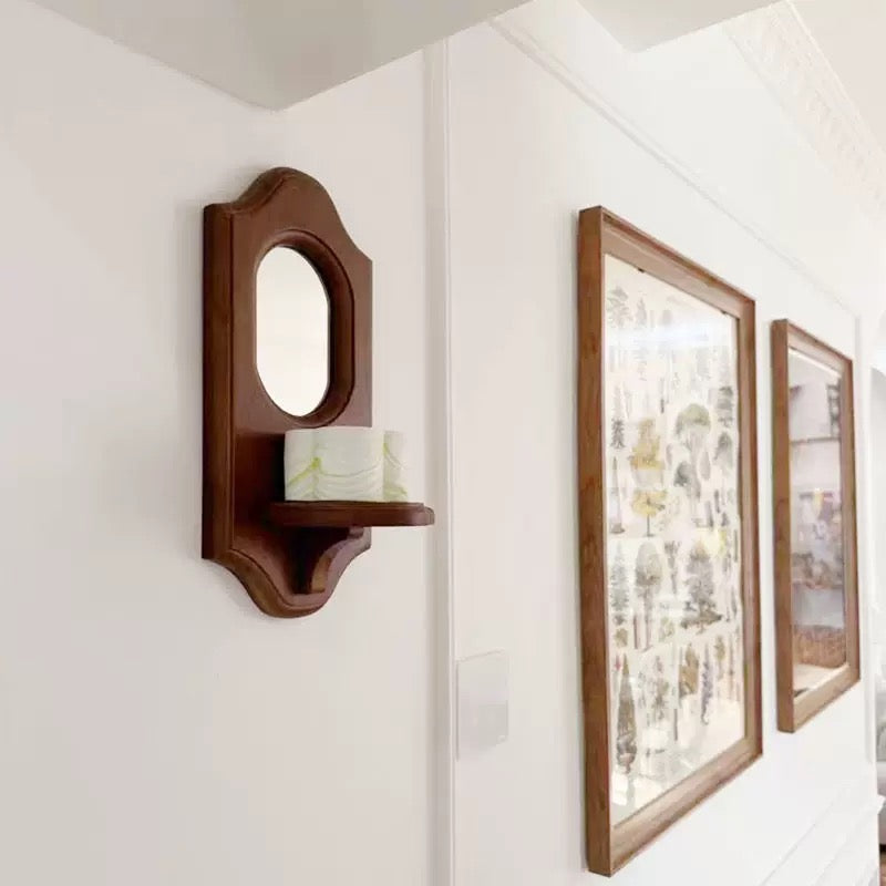 Wooden wall shelf with a mirror and candle next to framed artwork on a white wall.