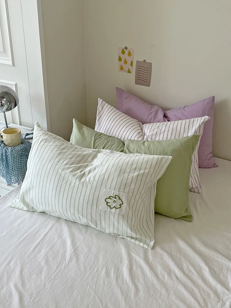 Sage flower embroidered pillowcase layered with lilac stripes and soft green pillows on a white bed.