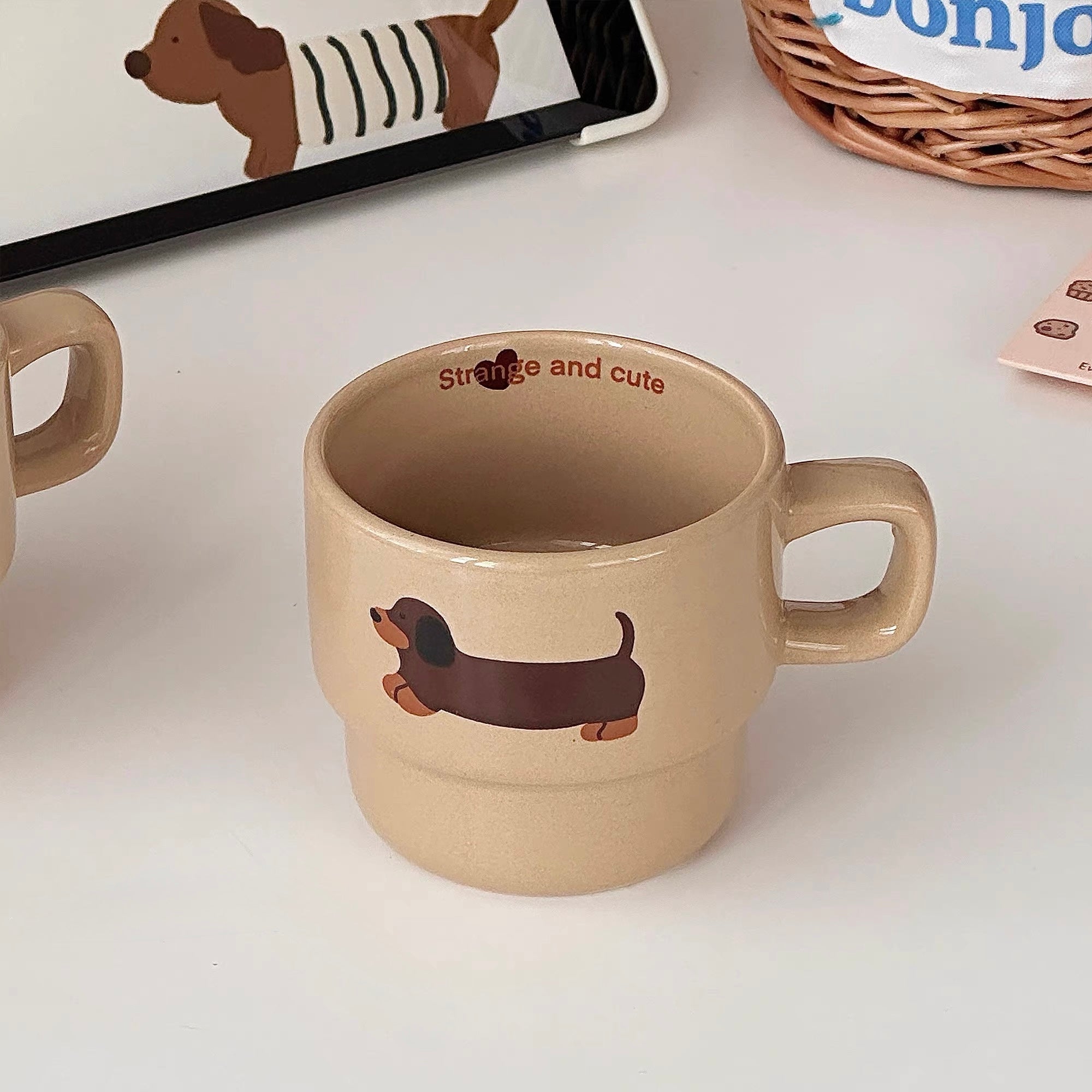 A beige ceramic mug with a dachshund illustration and the words 'strange and cute' printed inside, shown on a white surface with partial props around it.
