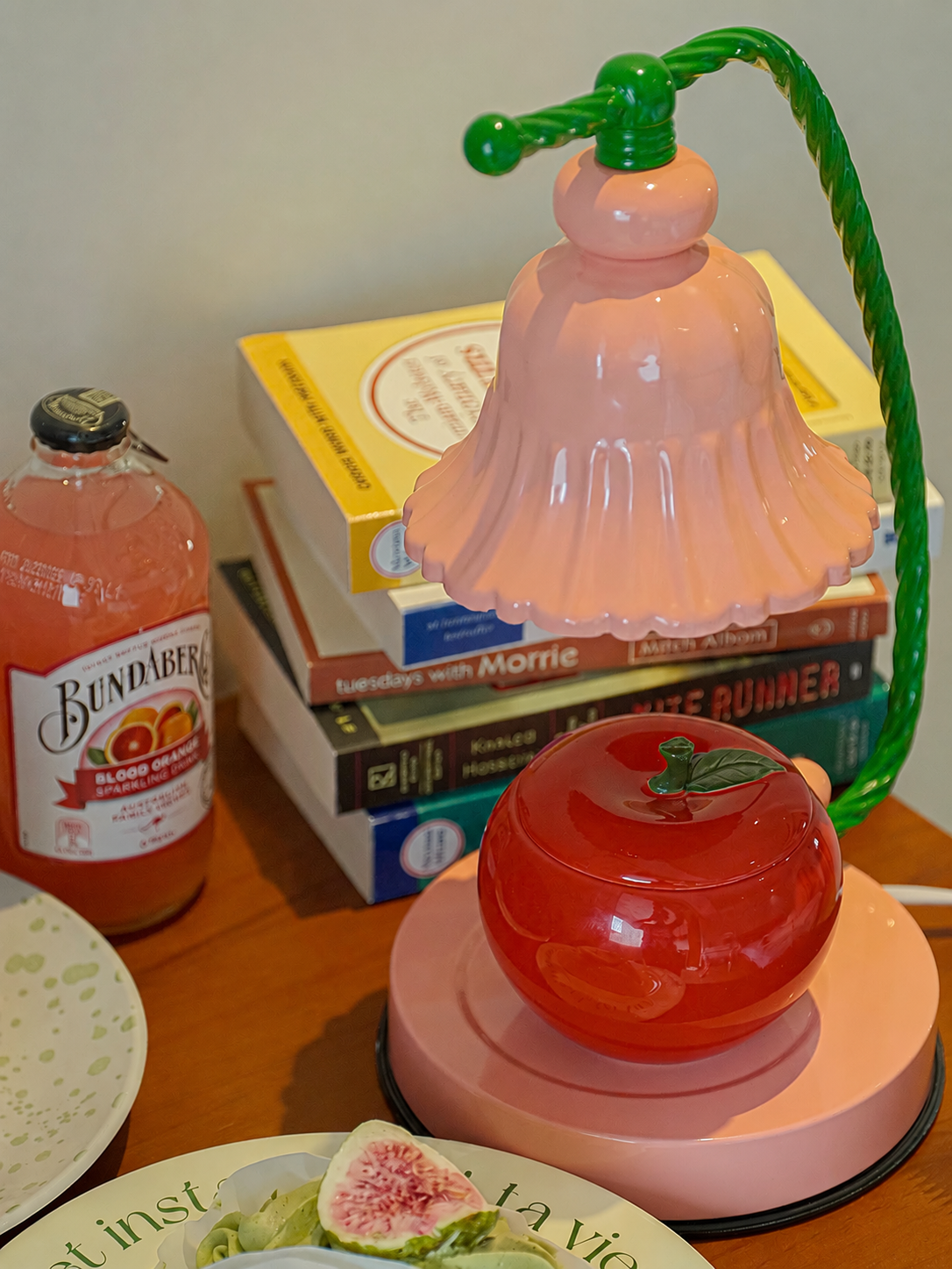 A pink bell-shaped table lamp with a green twisted cord and stem, featuring a glossy red apple-shaped shade or container on a matching pink base, set on a wooden table with books and a bottle in the background.