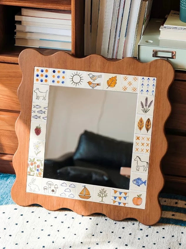 A scalloped mirror with a brown outer edge and illustrated border hangs above a cabinet with an open book and tea set.