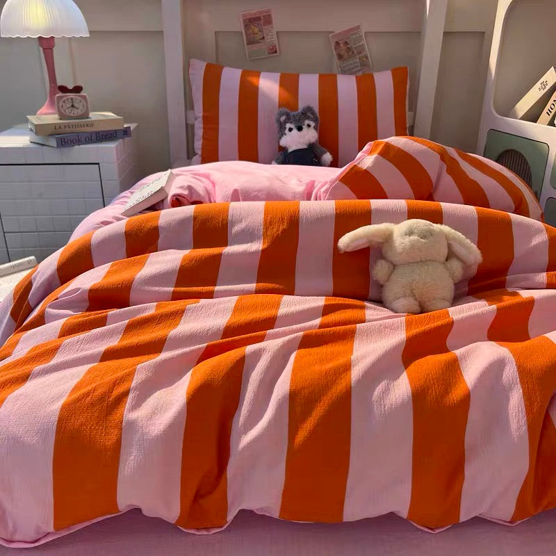 A cozy bed styled with orange and white striped bedding, including a duvet cover and pillowcase, with two plush bunny toys resting on the bed. The background shows a white wooden headboard and a bedside table with a lamp.