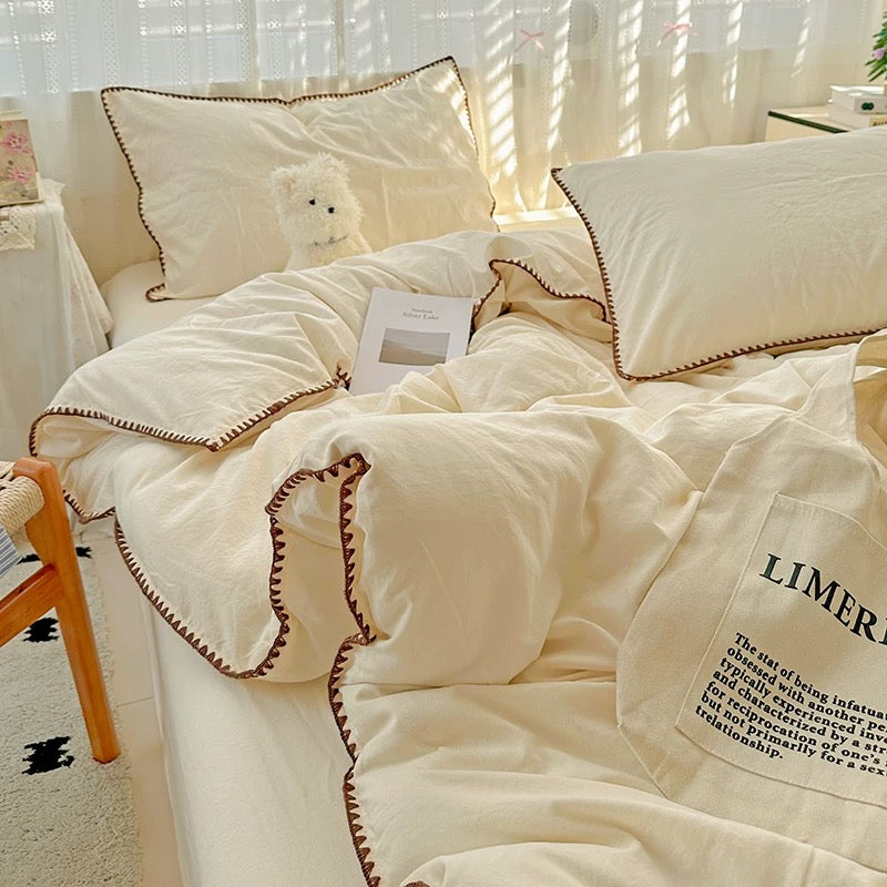 Cream-colored bedding set with brown contrast stitching and a scalloped edge, styled on a bed in a softly lit bedroom with sheer curtains and a small chair visible.