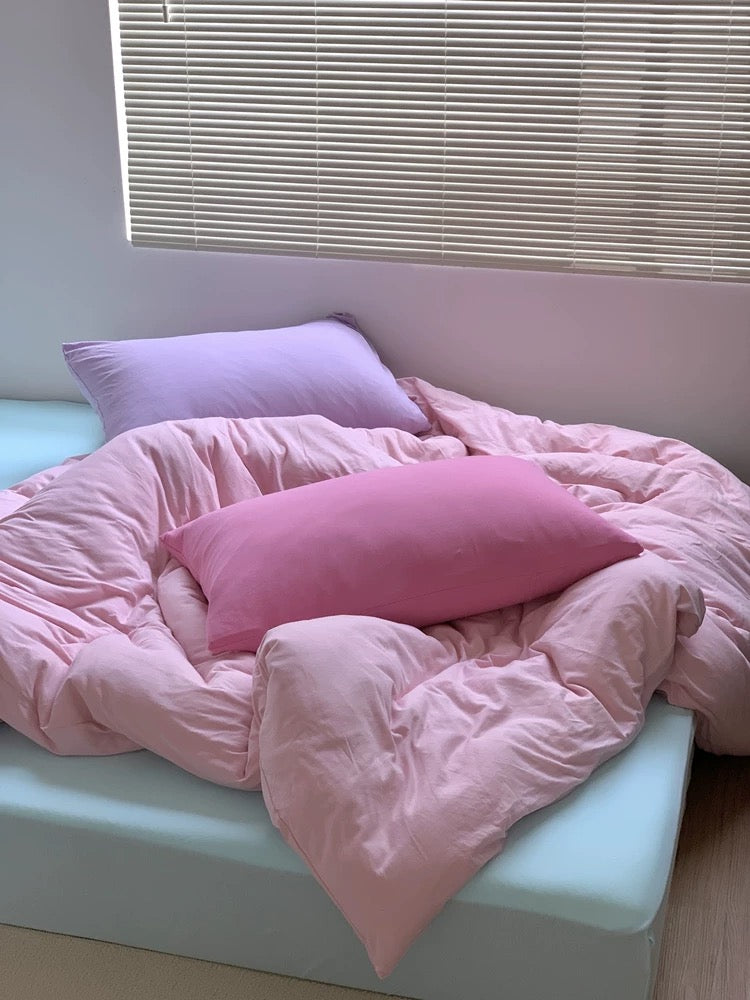 Pink comforter and pillows on a bed with light-colored walls and window blinds.