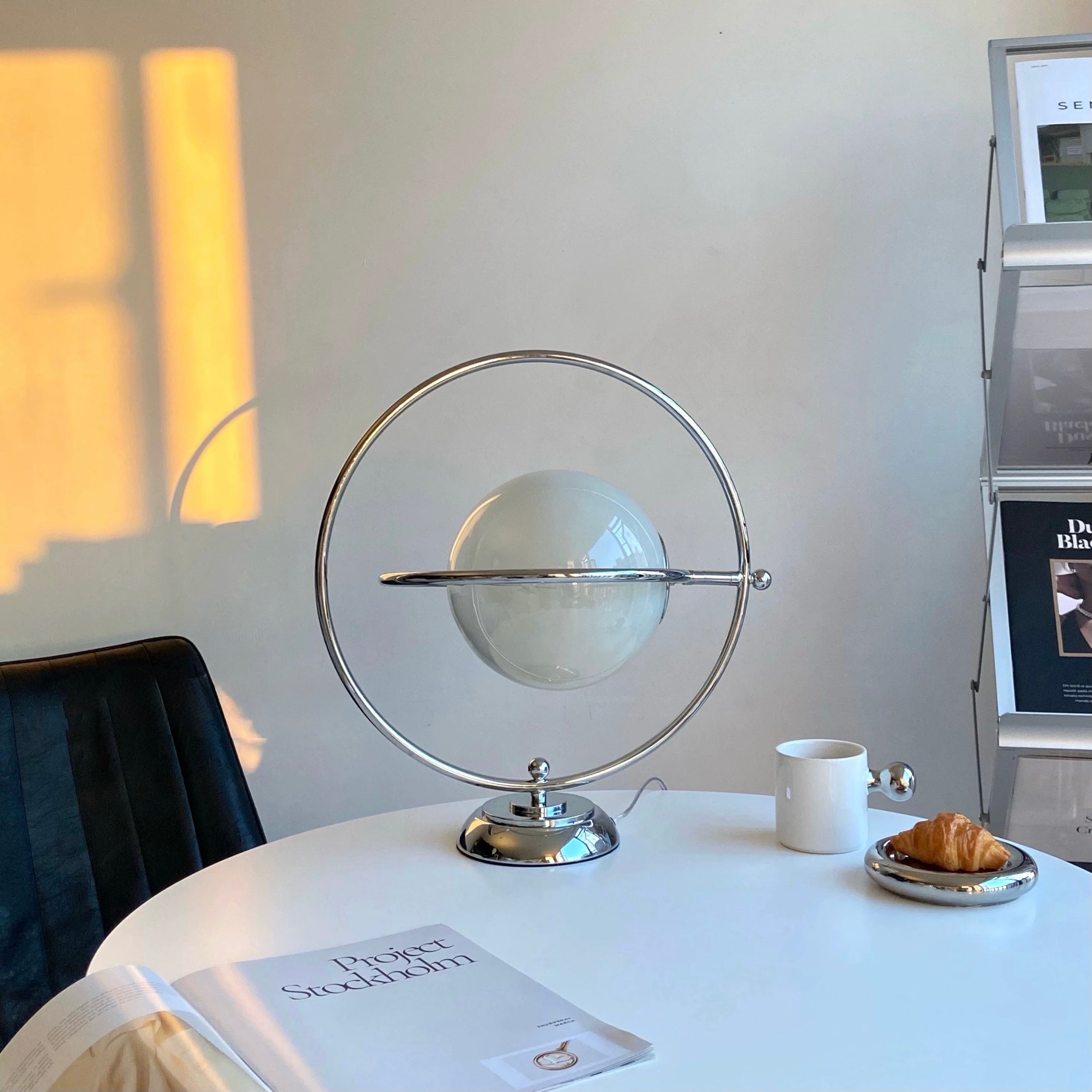 A modern spherical table lamp with a clear glass globe and silver metal frame, displayed on a white round table in a bright room next to a white mug and a croissant.