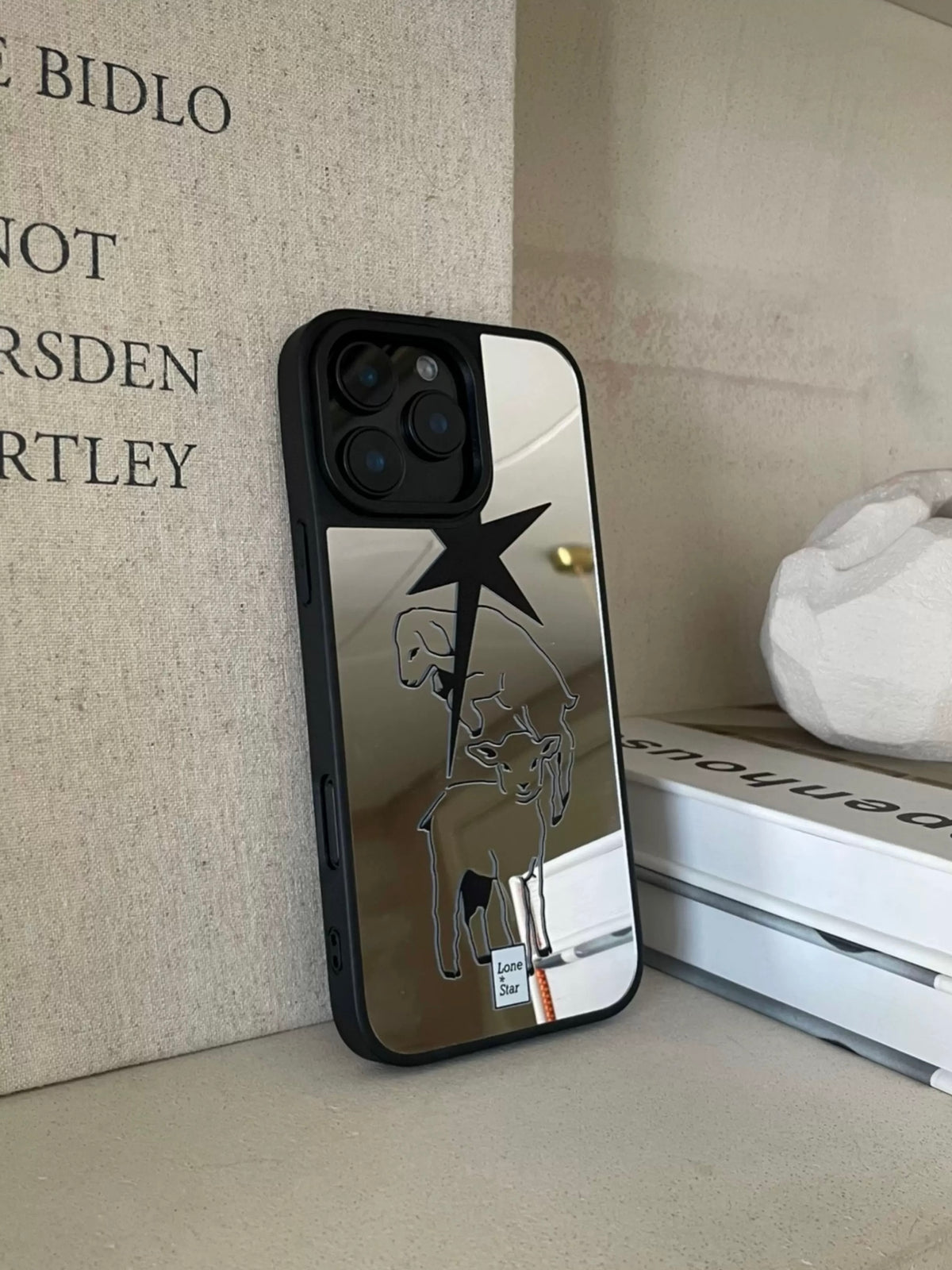 A silver mirror phone case stands upright with a large black pointed star above lamb outlines and black edging.