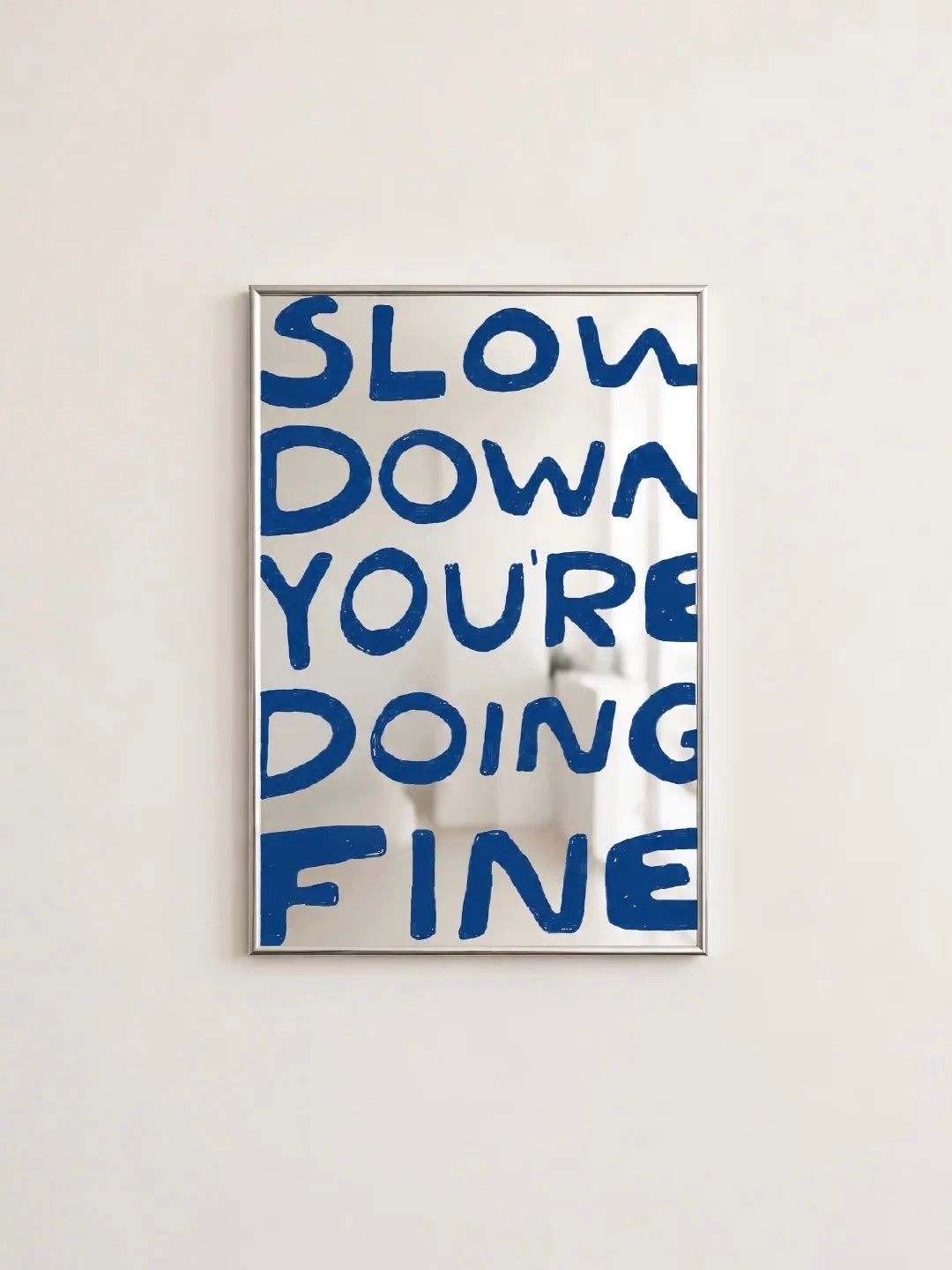 The Slow Down lettering mirror shows large blue handwritten words across its surface inside a slim silver frame.