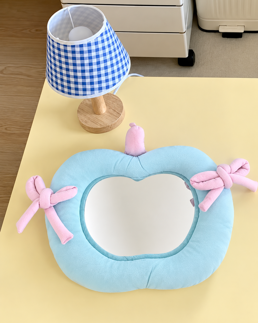 A light blue pumpkin-shaped wall mirror with pink bows and a small pink top ornament, displayed on a white wall above a styled white counter with a teddy bear, coffee machine, baskets, and small decor items.