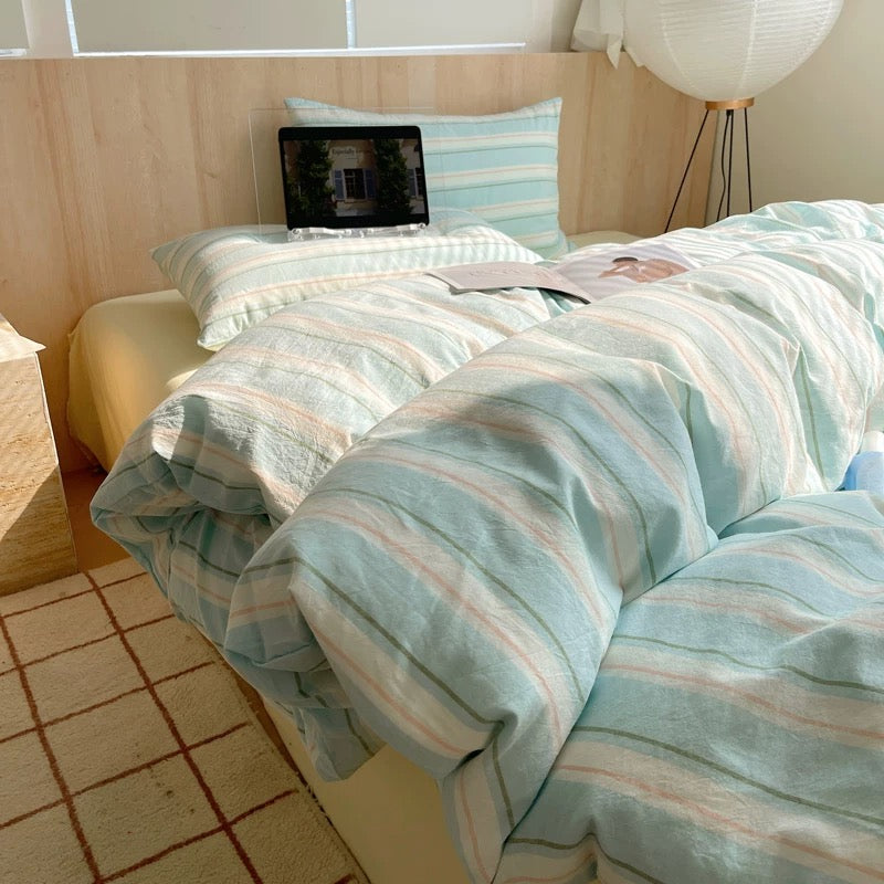 Bed with striped bedding and a laptop on a pillow, in a room with a lamp and window.