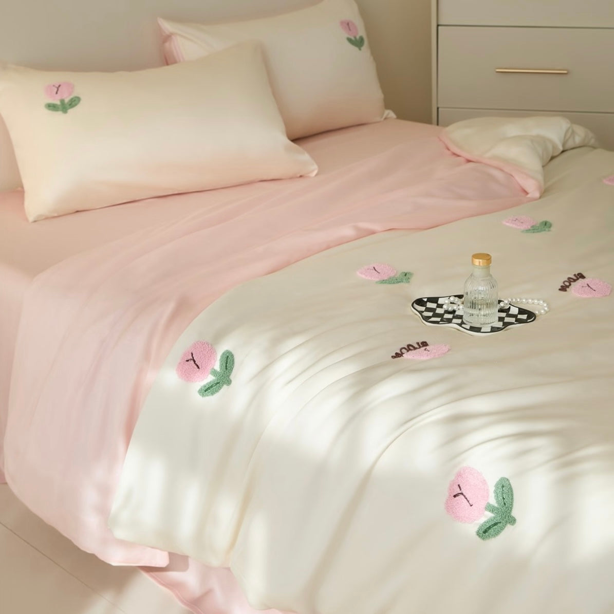 A neatly made bed with pale pink and cream bedding, decorated with embroidered pink flowers and green leaves. A small glass bottle sits on a black-and-white checkered coaster on the duvet.