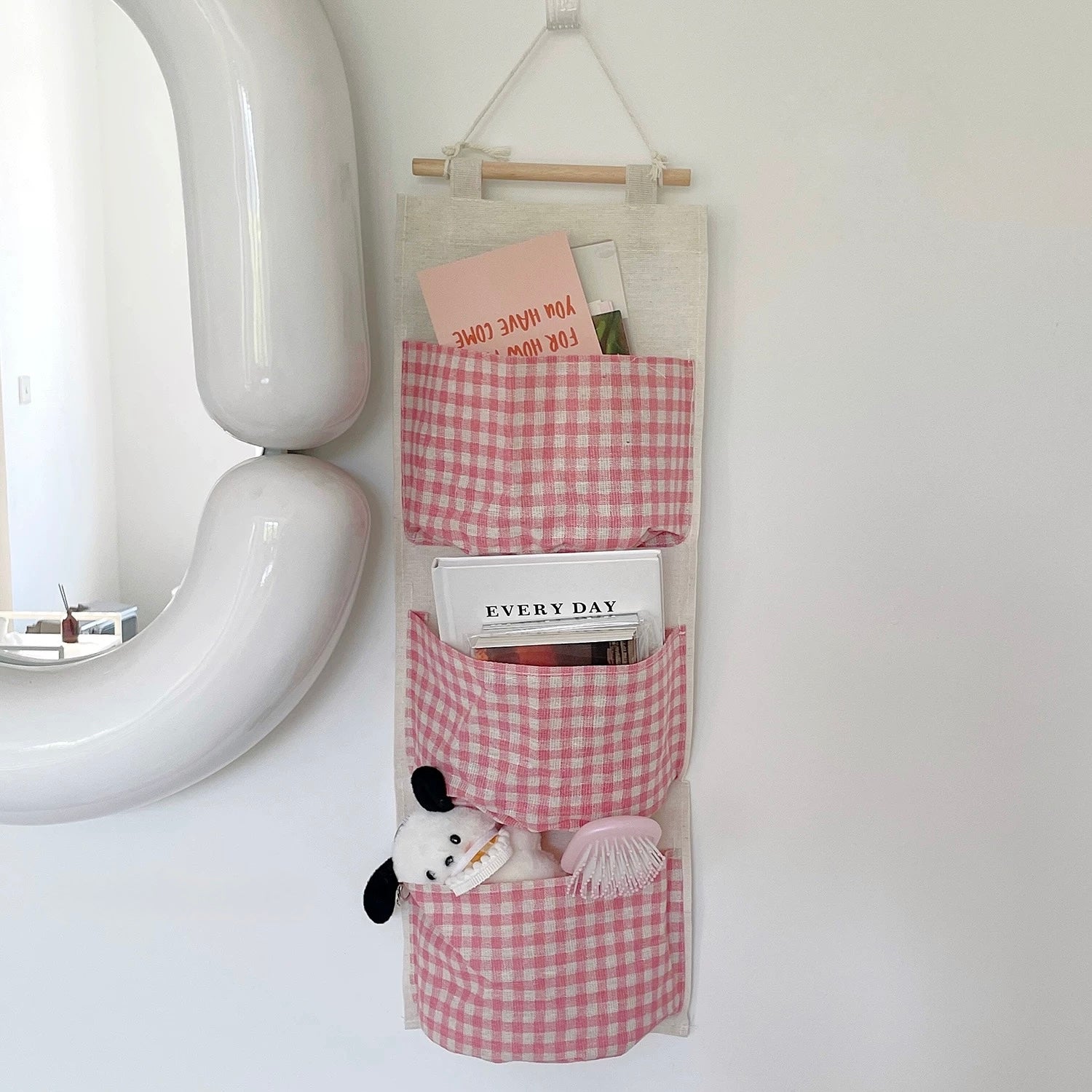 Pink gingham hanging organizer with books and a plush toy on a white wall.