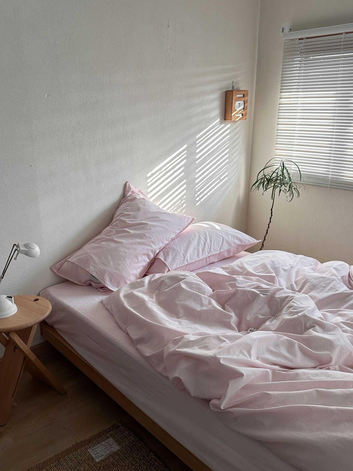 A bed dressed in light pink bedding with two matching pillows, styled in a minimalist bedroom with a wooden side table, lamp, window blinds, and a small plant.