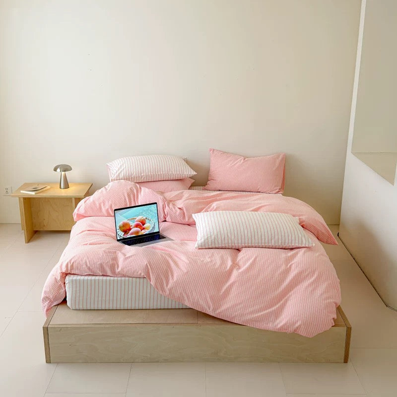 A bed dressed in a light pink and white striped bedding set, including a duvet cover, two pink pillows, and two striped pillows, set in a minimalist room with a wooden platform bed and a small side table.