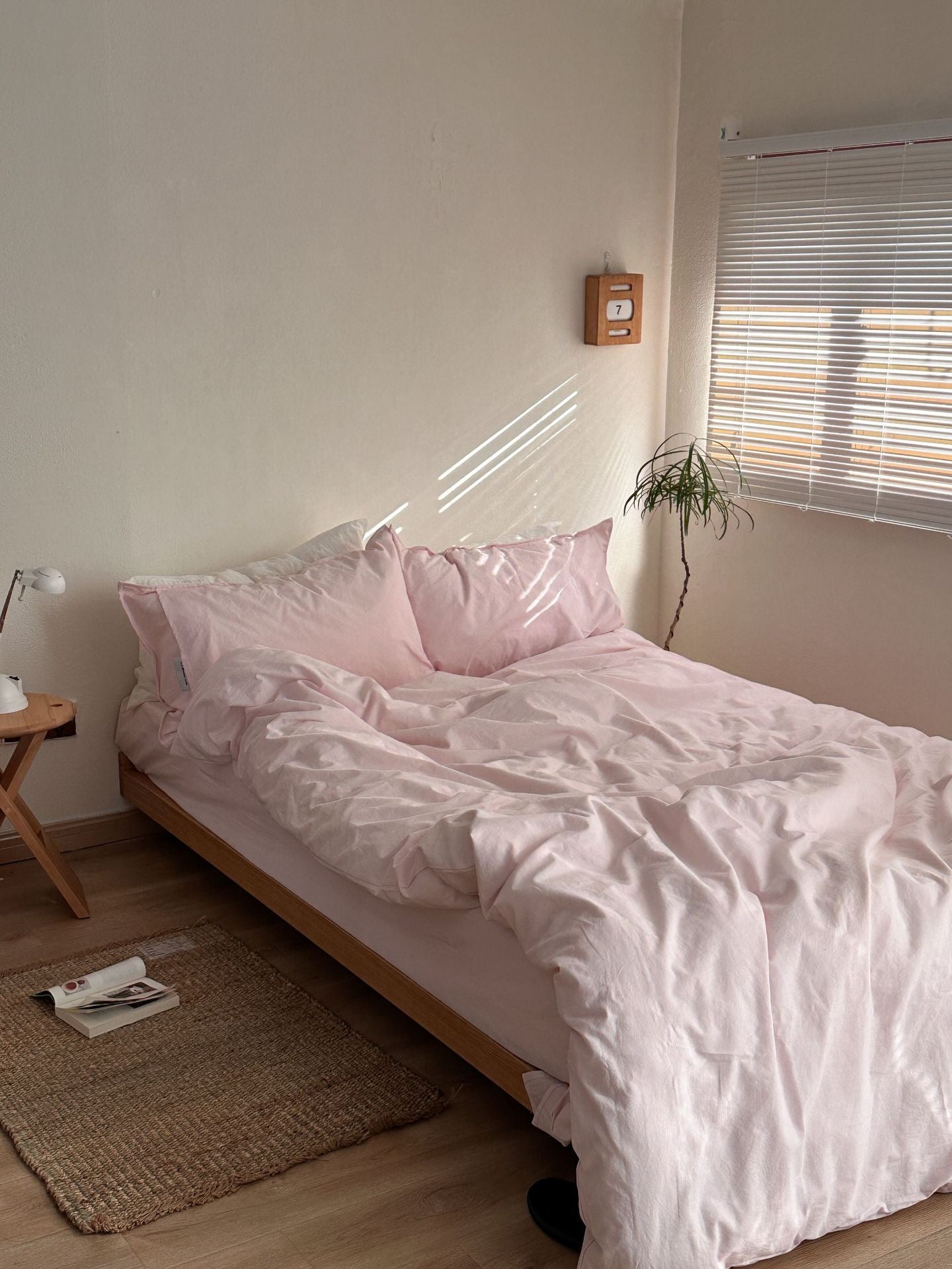 A neatly made bed in a minimalist bedroom setting, featuring light pink bedding with matching pillows and a duvet cover. The bed has a light wood frame and is positioned next to a small wooden side table with a lamp, a woven rug on the wooden floor, and a small potted plant near the window with blinds.