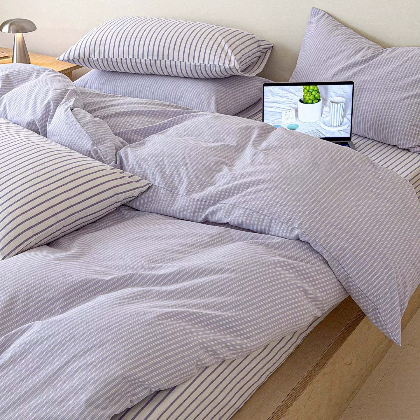 A bed dressed in light lavender and white striped bedding, including a duvet cover and matching pillows, with a small laptop resting on the bed.