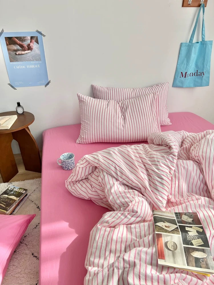 Pink bed with striped bedding in a room with a small table and lamp.