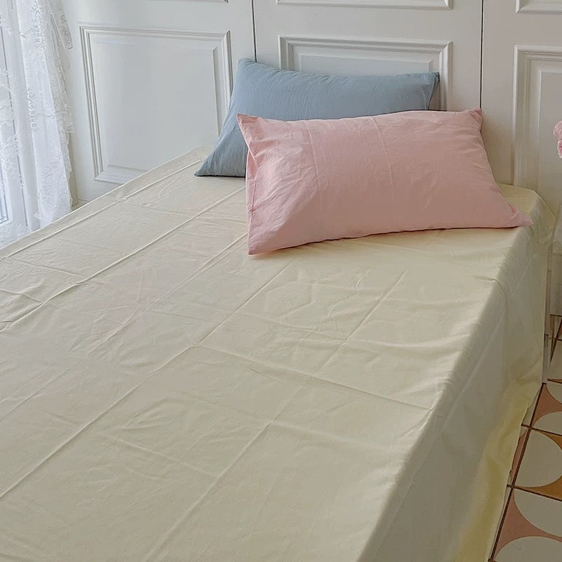 A cream-yellow sheet is paired with blue and pink pillows beside a sheer-curtained window.