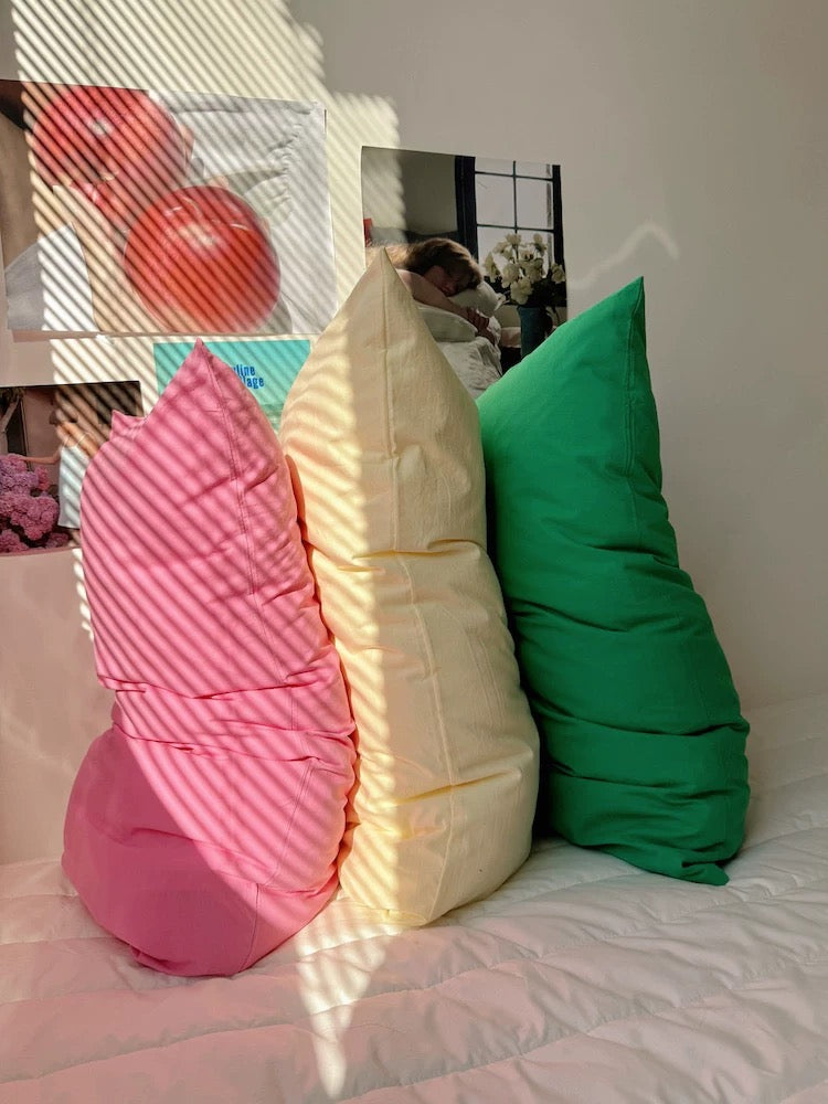 Three oversized decorative pillows in pink, cream, and green, styled on a white quilted bed with warm sunlight and window shadows.