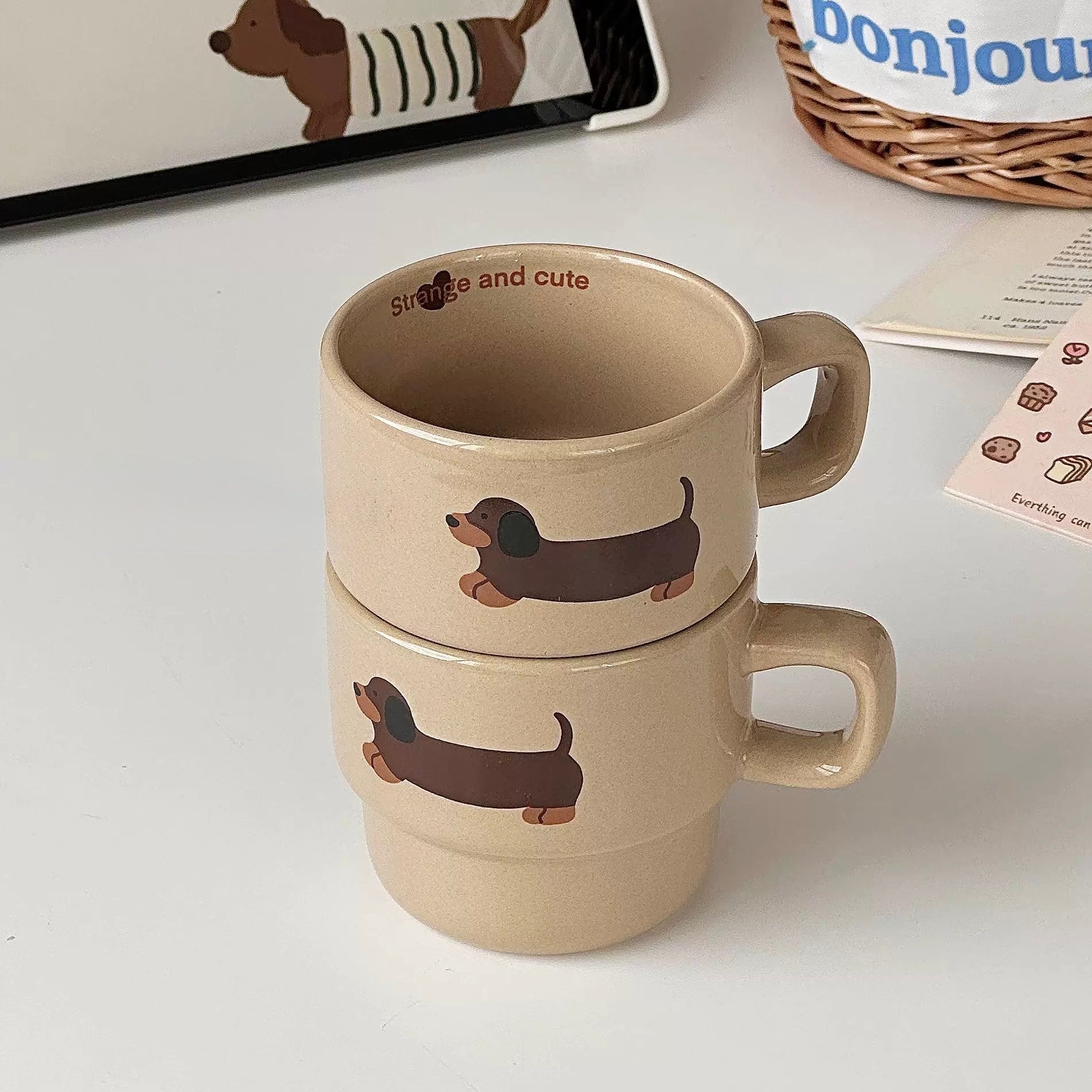 Two stacked ceramic mugs in a warm beige tone, each printed with a dachshund illustration and the words 'smile and cute'. The mugs are shown on a white tabletop with a few styled props in the background.
