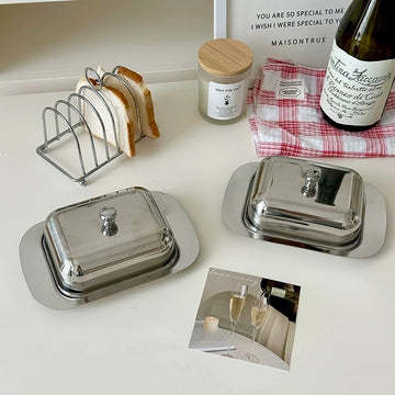 Silver butter dishes on a table with toast, candle, and bottle of wine.