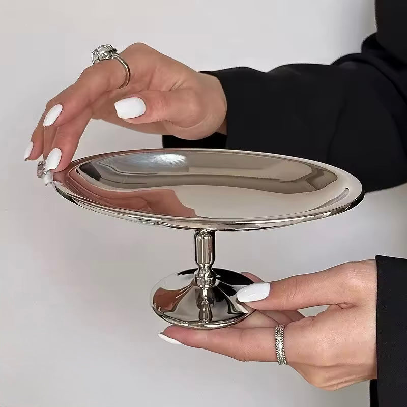 Silver cake stand held by a person with white nail polish against a plain background