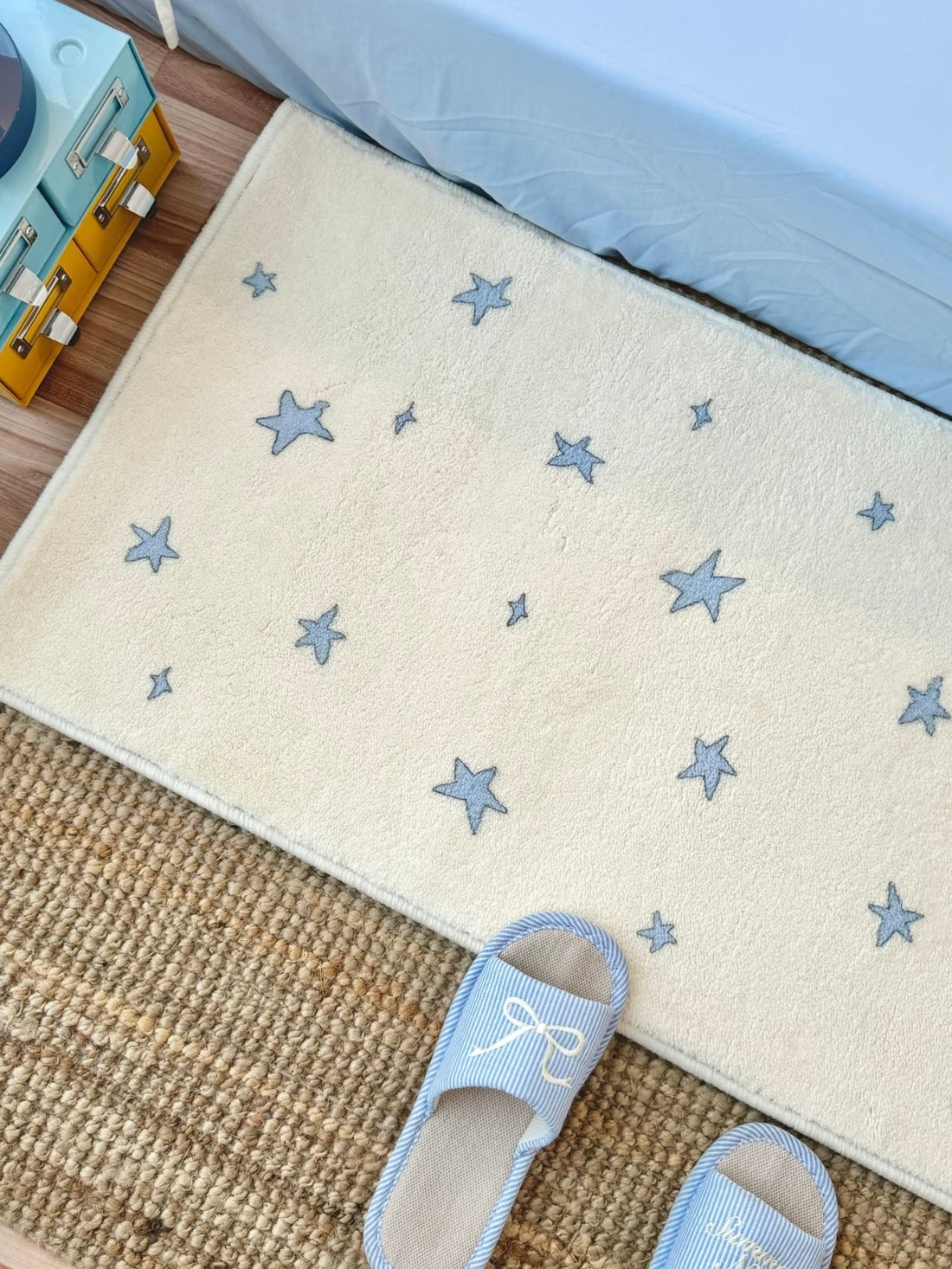 A star rug with blue motifs on a cream background lies beside a blue bed, with slippers at the near edge.