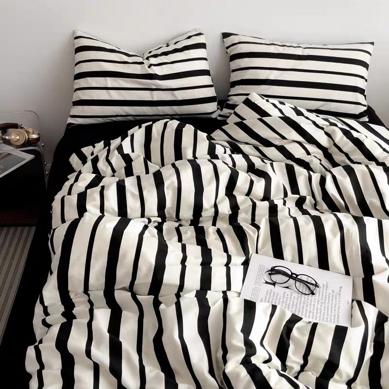Black and white striped bedding set with two matching pillowcases and a striped duvet cover, styled on a bed with a book and glasses in a bedroom setting.