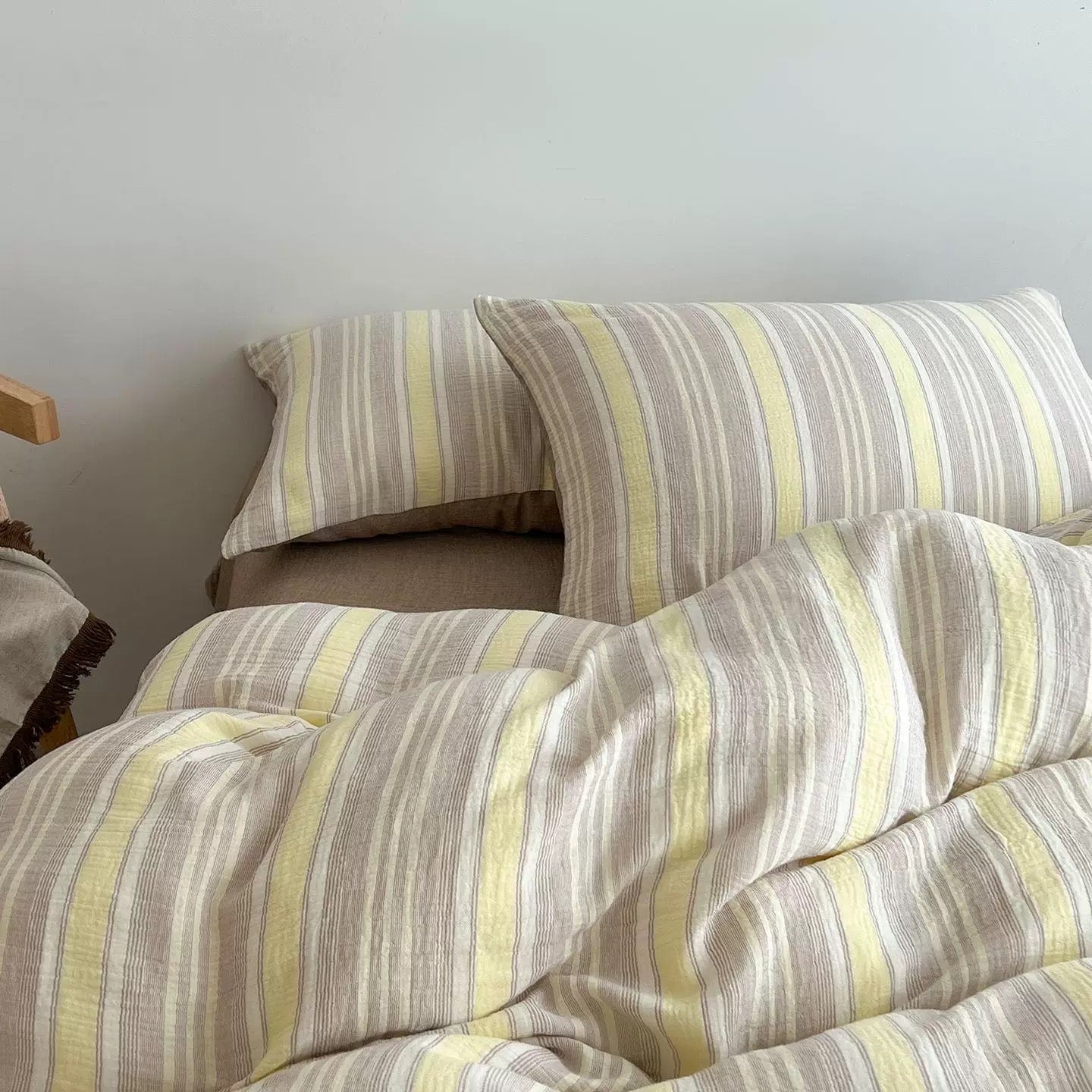 A close-up of a striped bedding set with a duvet cover and two pillowcases in a soft beige, gray, and pale yellow stripe pattern, styled on a bed against a plain light gray wall.