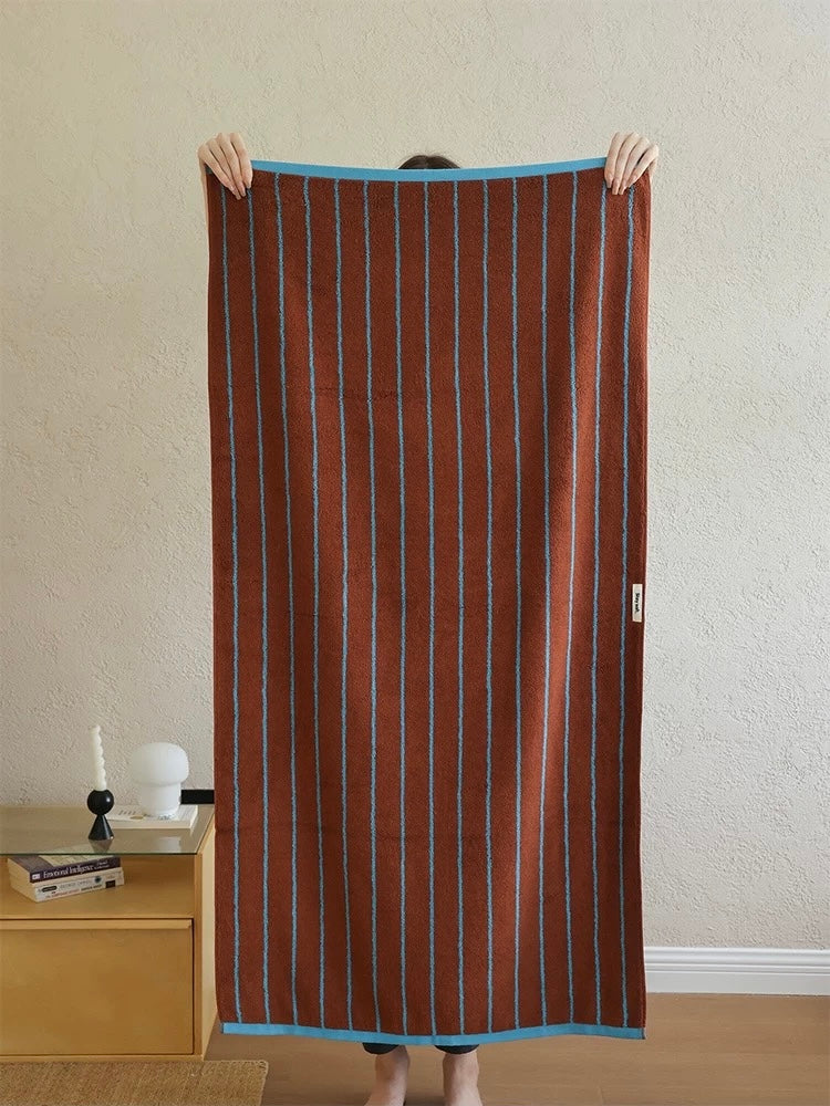 A person holds up a large rectangular brown towel with vertical light blue stripes and solid blue borders, standing in a room with a textured beige wall and a wooden side table.