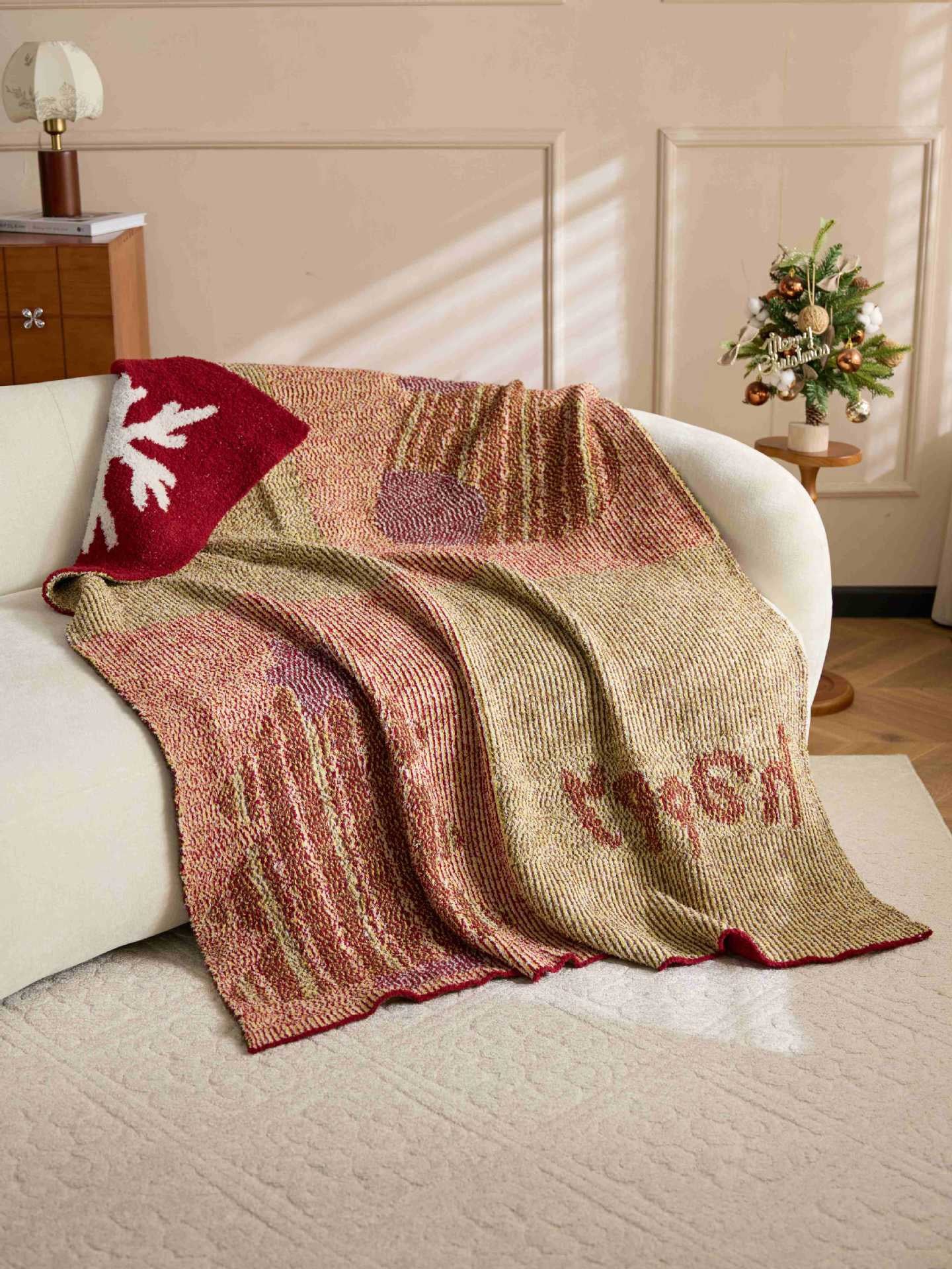 A knitted throw blanket draped over a white sofa in a warm, styled room. The blanket features a textured pattern of beige, rust red, and dusty pink, with a solid red section on one end featuring a white snowflake motif.