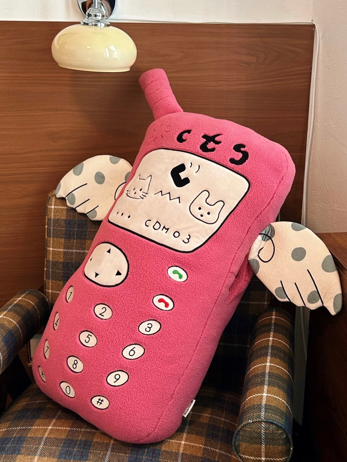 A pink telephone plush leans against a checked chair, showing its embroidered number buttons and spotted arms.