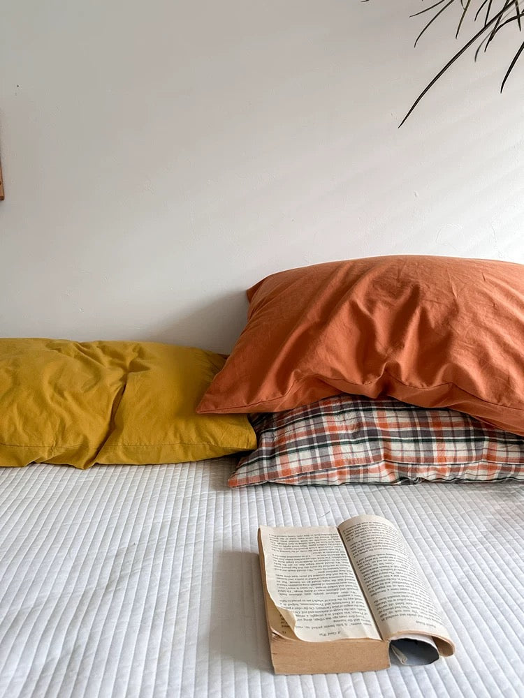An orange terracotta pillowcase rests above the plaid design beside mustard yellow on white bedding.