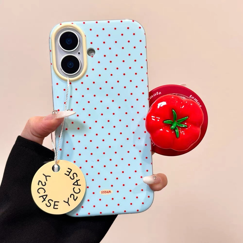 Phone case with red polka dots held by a hand, with a small red object and yellow tag in the background.