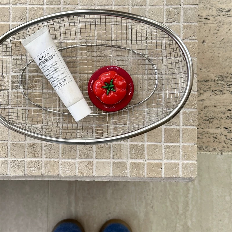 Metal strainer with a tube of cream and a red tomato-shaped object on a tiled surface.