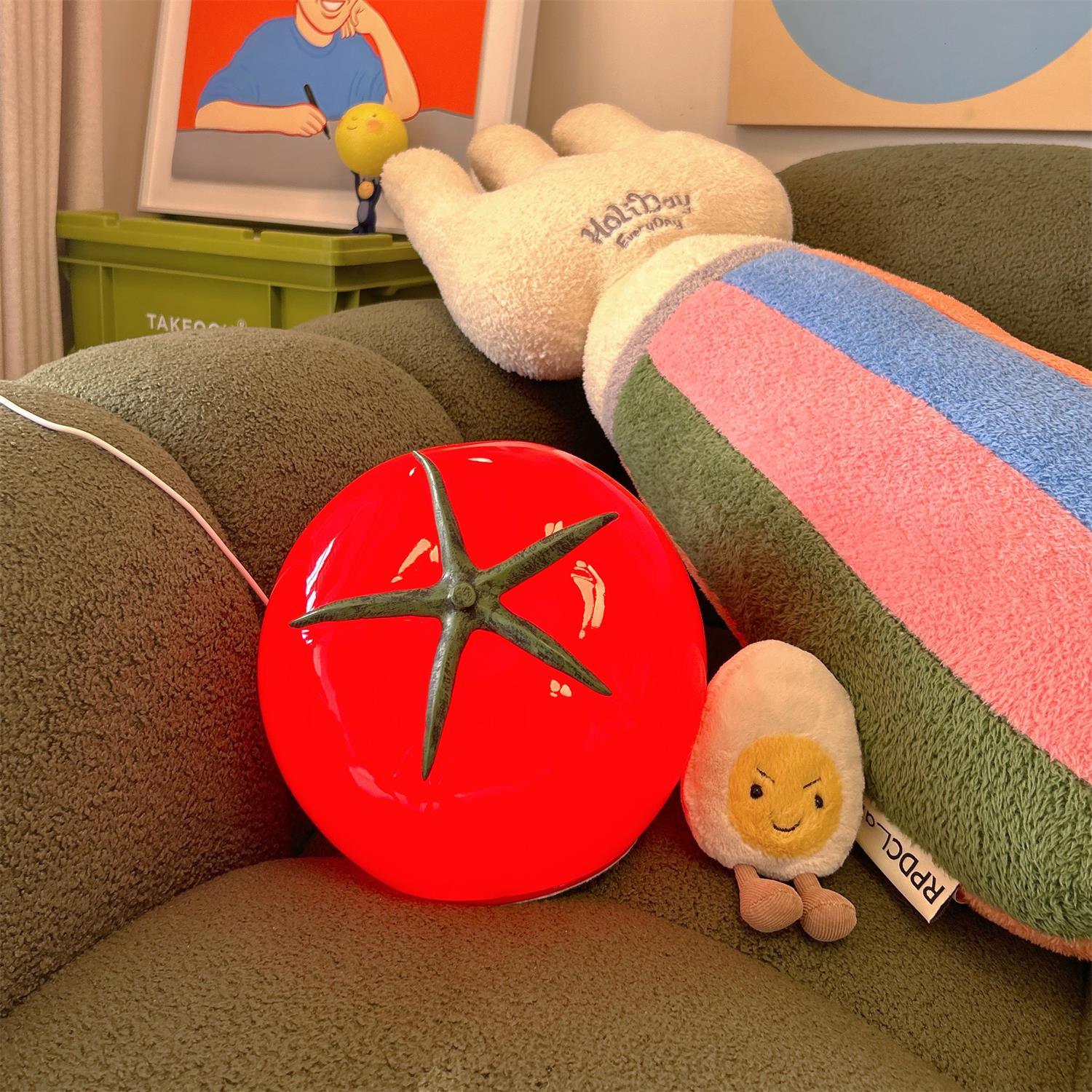 Plush toy tomato and egg on a couch with colorful cushions and a framed picture in the background.
